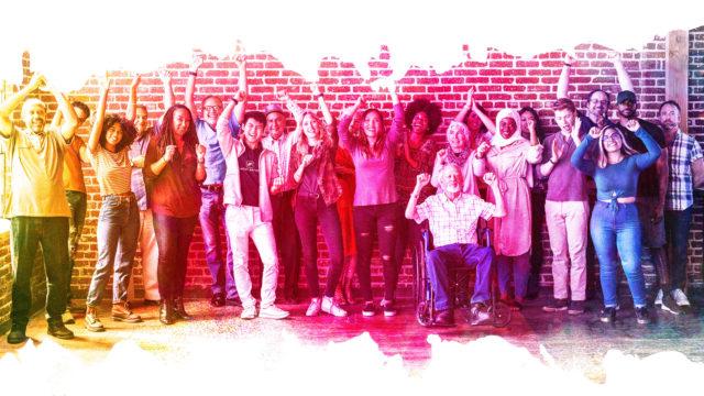 A diverse group of people stand against a brick wall with a transparent rainbow colour overlay